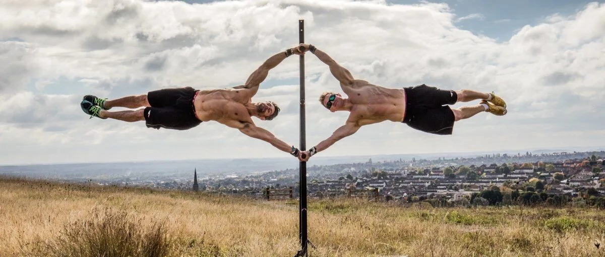 Two athletes performing a synchronized human flag pose on a vertical pole outdoors